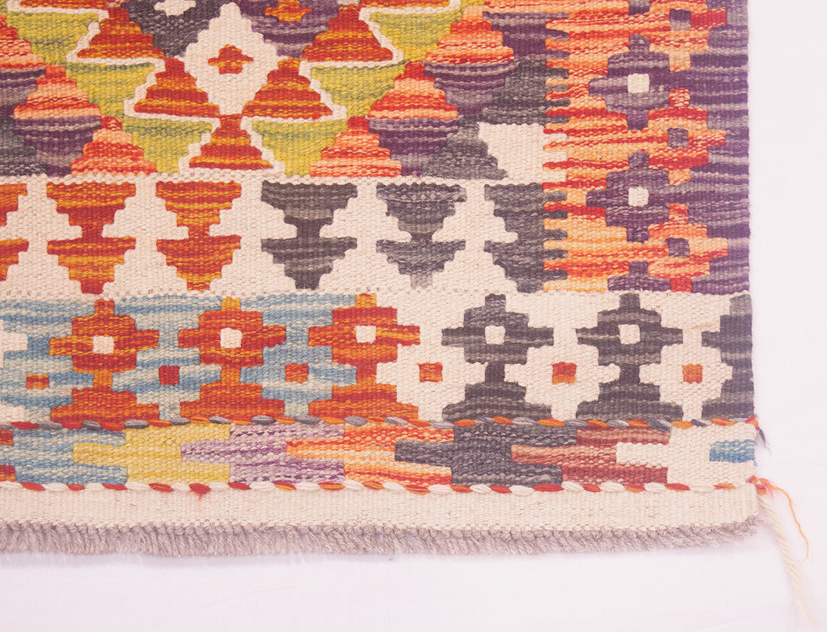 A close-up of a corner of a handwoven Afghan Kilim strip with a multicolour geometric pattern on a cream background, showing rows of orange, purple, grey, yellow, and blue motifs with a fringed edge.