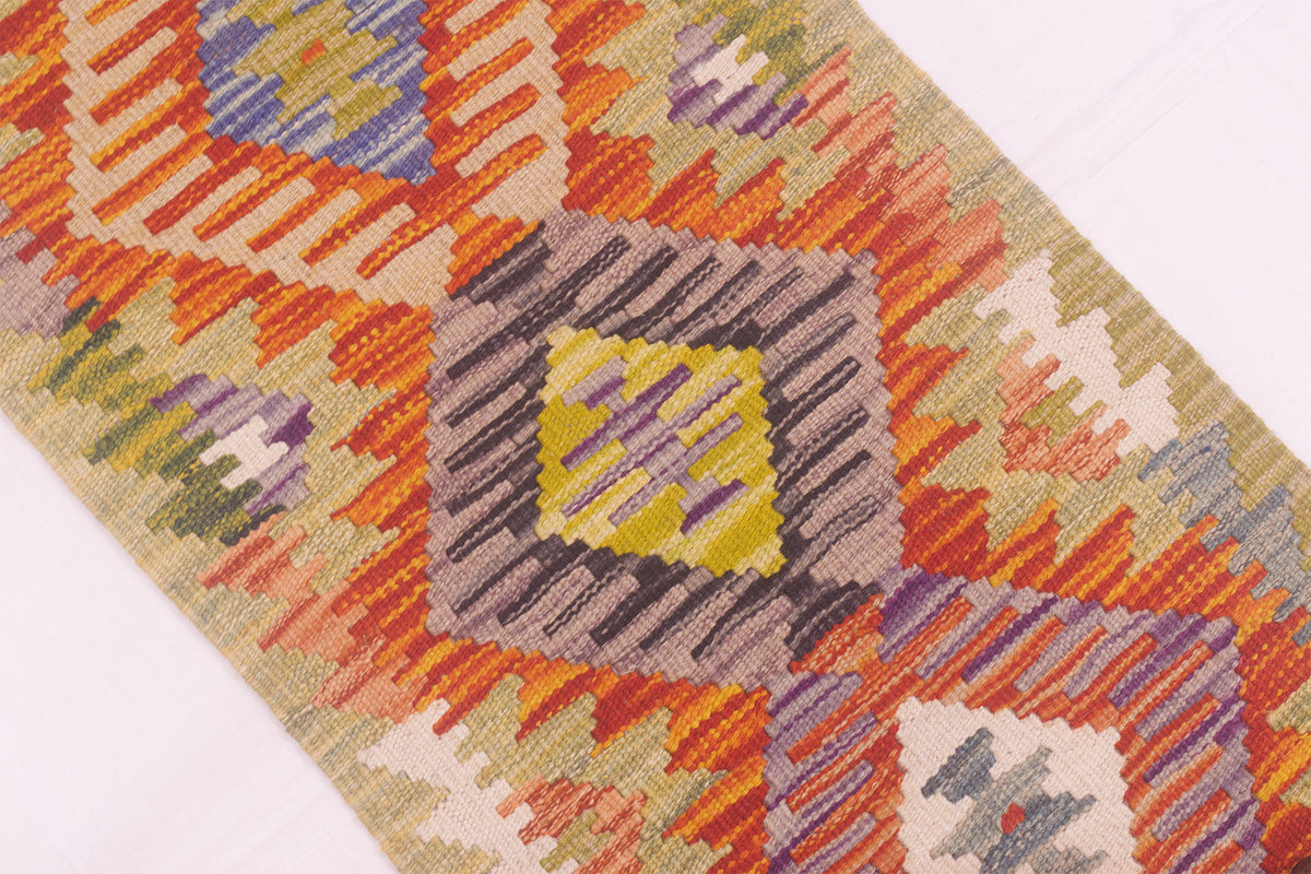A close-up of a woven kilim strip/runner rug with a geometric diamond pattern in orange, beige, purple, yellow, and green on a white background.