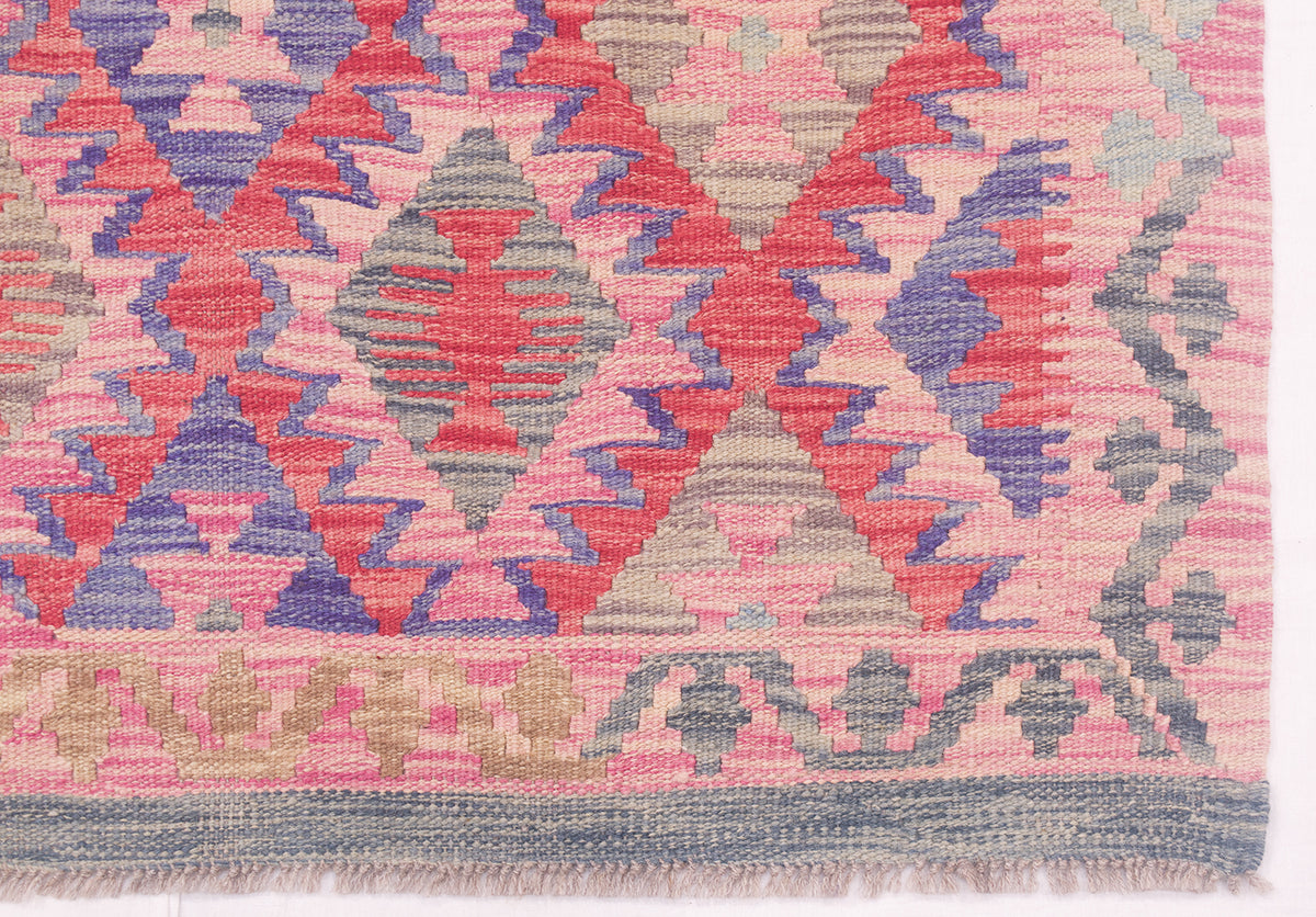 A close-up of a woven kilim Afghan rug showing a geometric diamond pattern in pink, coral, purple, and grey, with a fringed edge at the bottom.