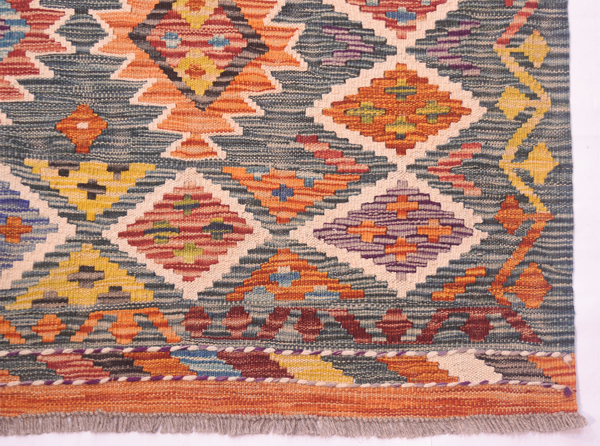 A close-up of a handwoven geometric Kilim Afghan rug showing a tribal pattern with orange, yellow, red, and purple diamonds on a green background, finished with a fringed edge.
