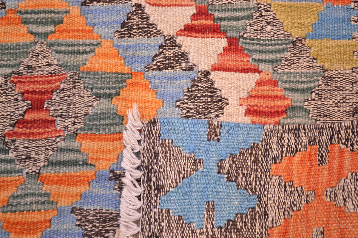 A close-up of the handwoven back of a woven Afghan kilim rug featuring a geometric pattern of triangles and diamonds in orange, blue, red, beige, grey, and green, with a white fringe visible on the left.