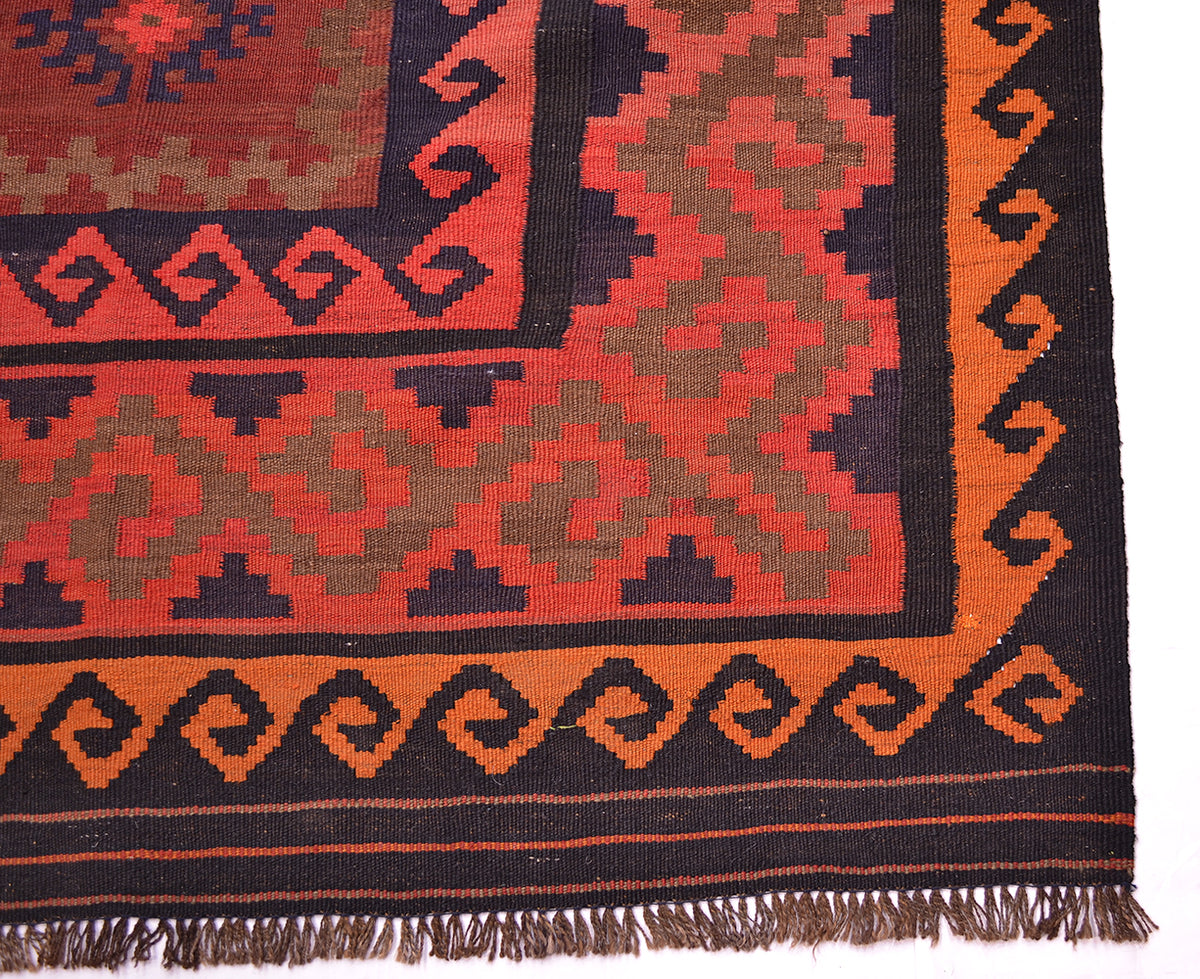 Close-up of a patterned old Afghan kilim rug corner showing a geometric design in rust red, orange, olive green, and black with fringed edge.
