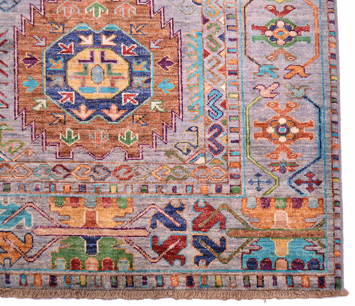 A close-up of a traditional geometric patterned Afghan Waziri rug with a central medallion, border motifs, and fringe at the bottom edge. The design features multiple colours including blue, orange, green, red, and beige on a light greyish field.