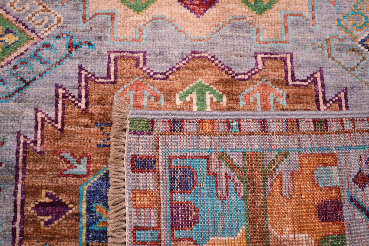 Close-up of a corner turned up on a handwoven Afghan Waziri rug corner showing a beige fringe, with a geometric pattern in muted blue, orange, purple, green, and brown on a light grey base.