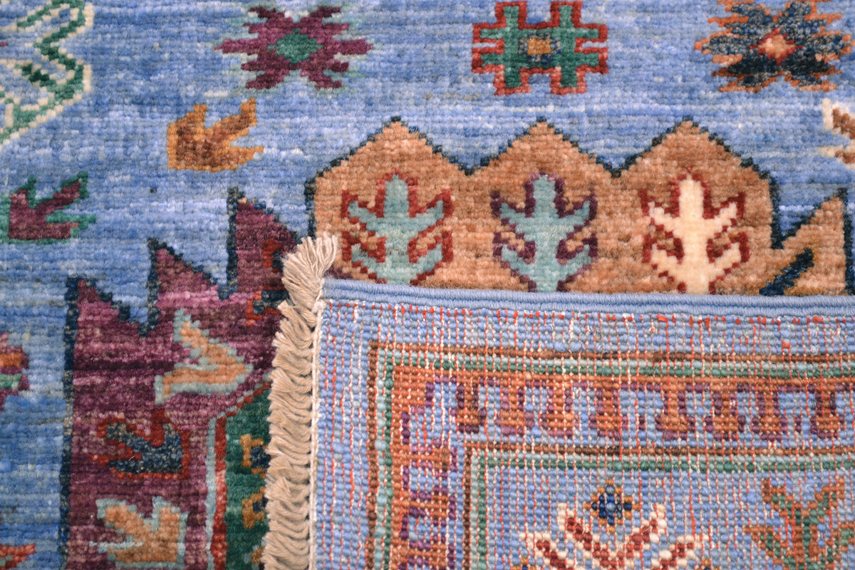 Close-up of a corner turned up revealing the hand-knotted weave of a handmade Waziri runner showing a light blue field with geometric and floral motifs, a tan border section, and cream fringe on the left edge.