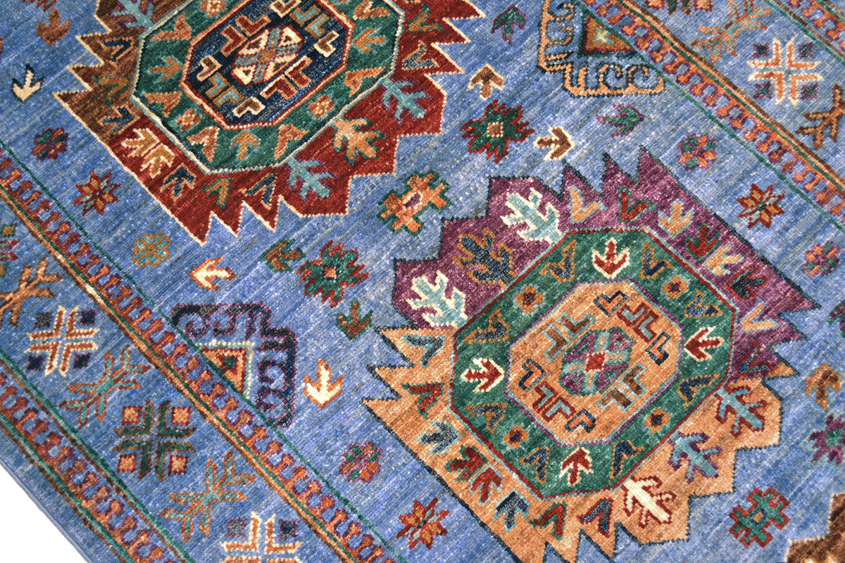 A close-up of a traditional patterned Afghan Wazir runner with a dusty blue field, featuring large central medallions in green, rust red, purple, and tan, surrounded by smaller floral motifs.