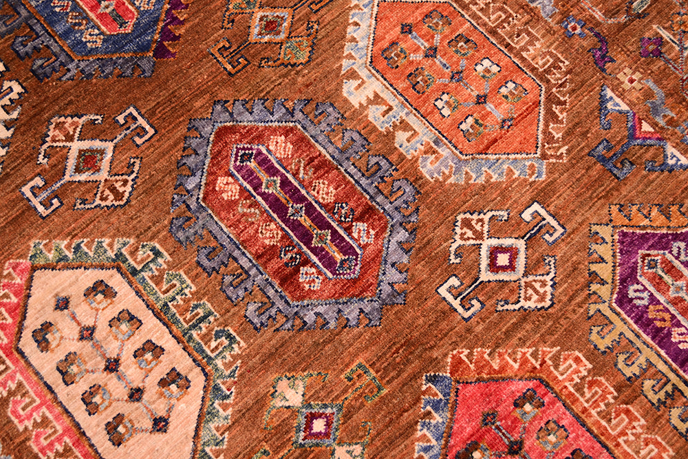 A close-up of a traditional patterned Afghan Waziri rug with a warm brown base and geometric motifs in red, orange, purple, beige, and blue.