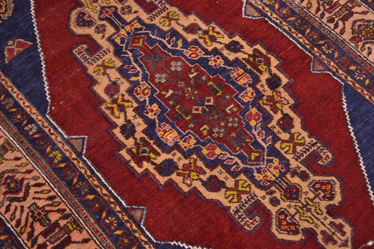 A close-up of a traditional handwoven old Turkish rug featuring a central geometric medallion pattern in deep red, navy blue, beige, and yellow.