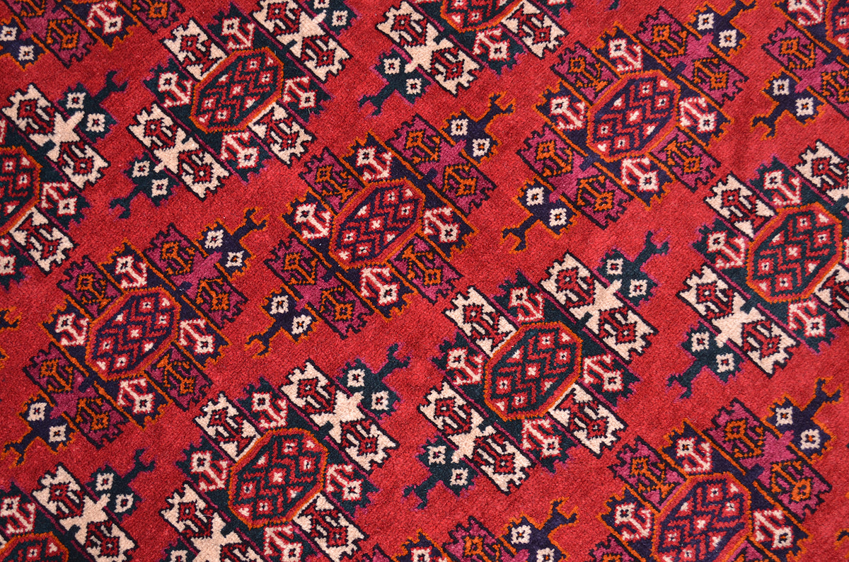 A close-up of a red patterned Yamoud rug with repeating geometric motifs in cream, navy, orange, and purple.