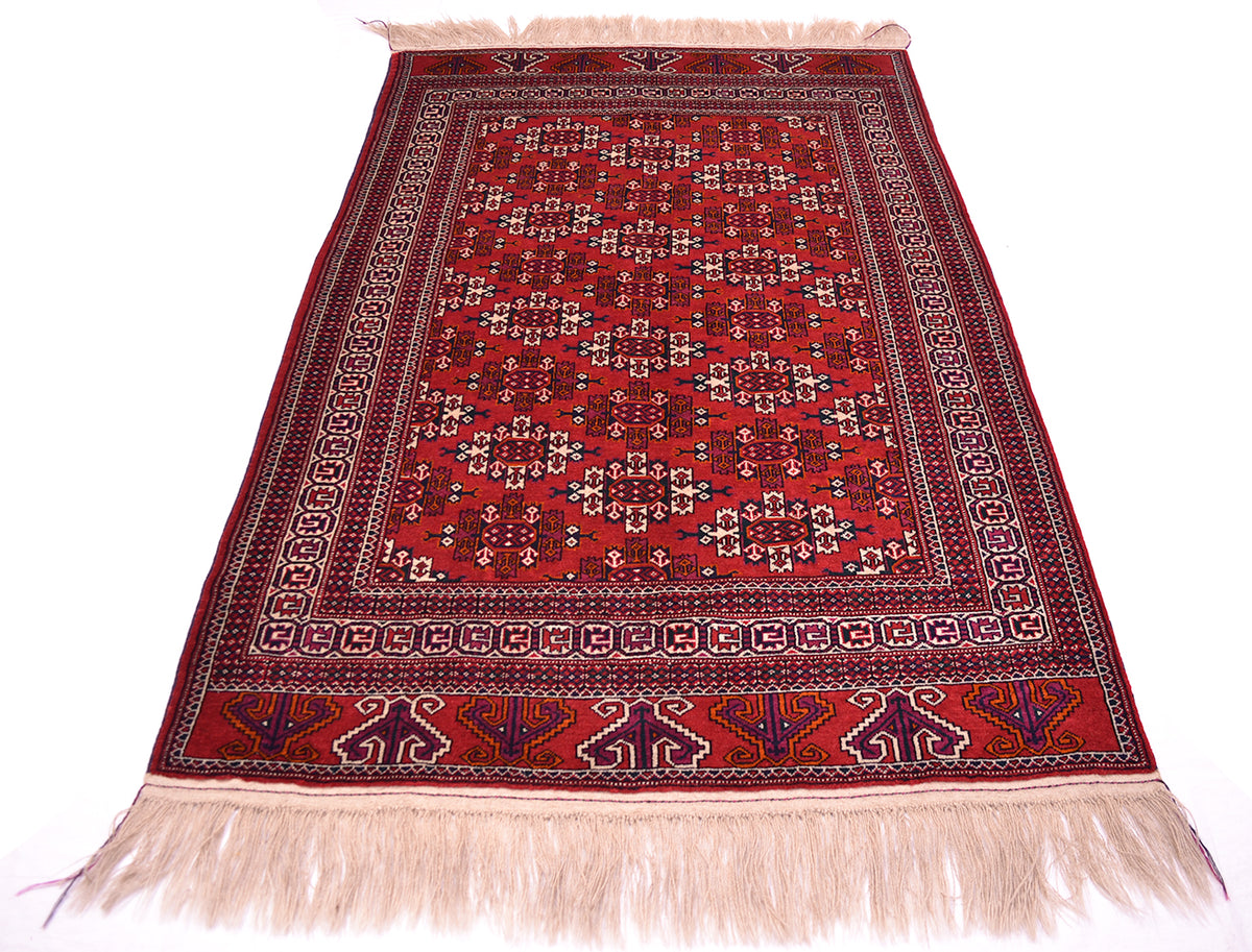 A traditional handmade red Turkoman wool rug with an intricate geometric pattern and cream-coloured fringe on the top and bottom edges, displayed flat against a white background.
