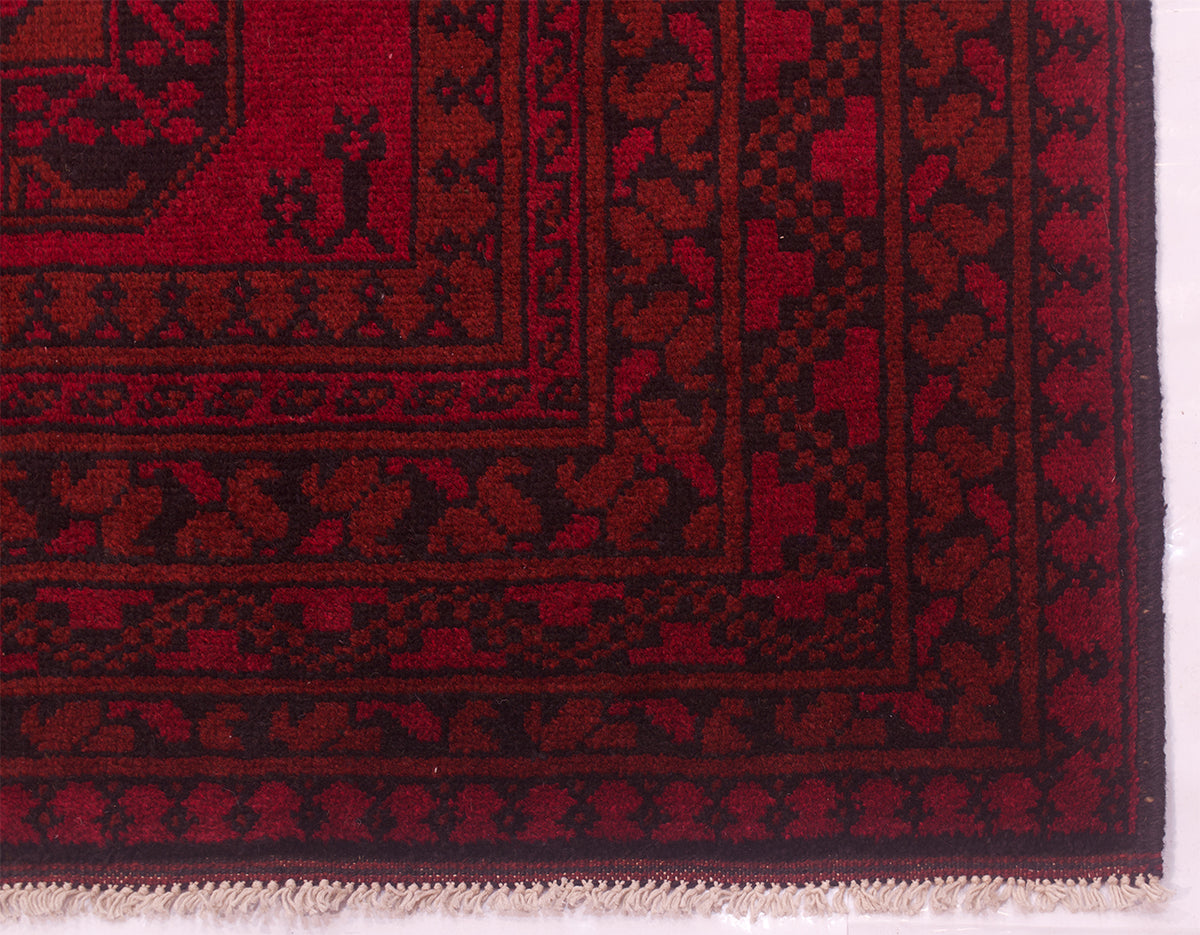 A close-up of a traditional patterned Afghan rug corner, showing deep red and black woven borders with a small white fringed edge.