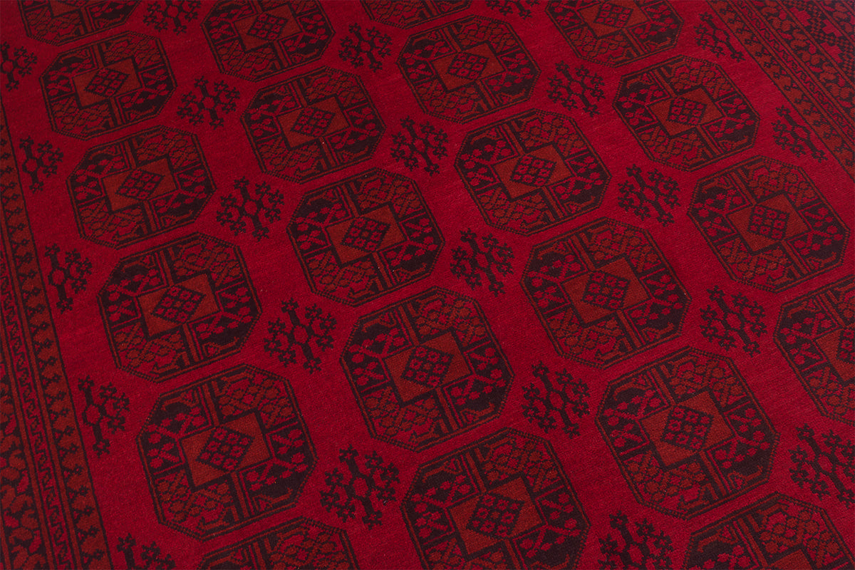 A close-up of a red Afghan Aqcha carpet featuring a repeating dark geometric pattern with octagonal medallions and small floral motifs.
