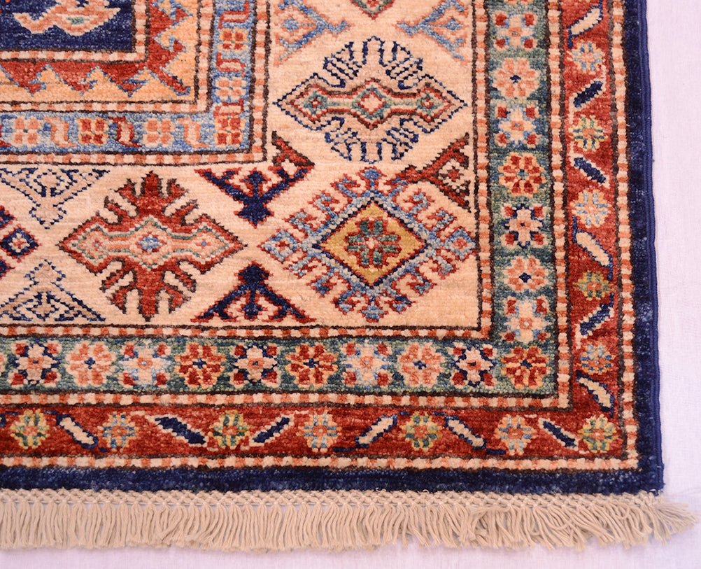 A close-up of a traditional handmade fine Kazak rug corner showing cream, rust red, navy blue, and teal geometric motifs with a fringed edge on a white background.