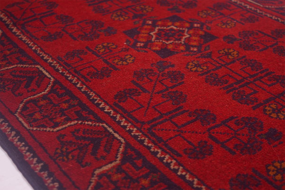 A close-up of a red patterned Afghan rug with dark navy and beige geometric motifs and borders.