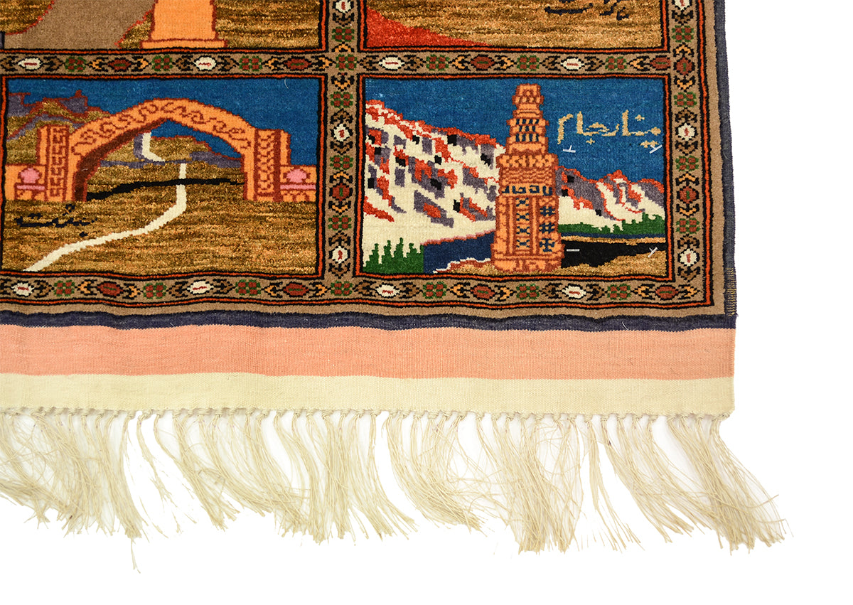 A close-up of a corner of a fine hand-knotted pictorial Afghan Kabul Mauri rug or mat corner showing a cream fringe, a peach border, and a multicoloured geometric and scenic pattern (showing Taq-e Zafar and the Minaret of Jam) with blue, red, green, and tan sections on a white background.