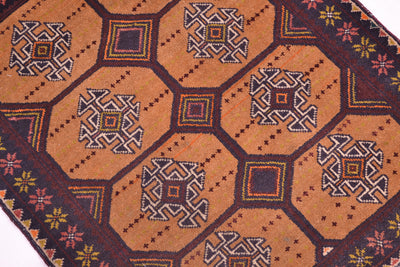A close-up of a patterned Old handmade Belouch mat / rug showing a warm fawn-brown base with repeating geometric motifs in dark brown, black, cream, and small accents of red, yellow, and blue.