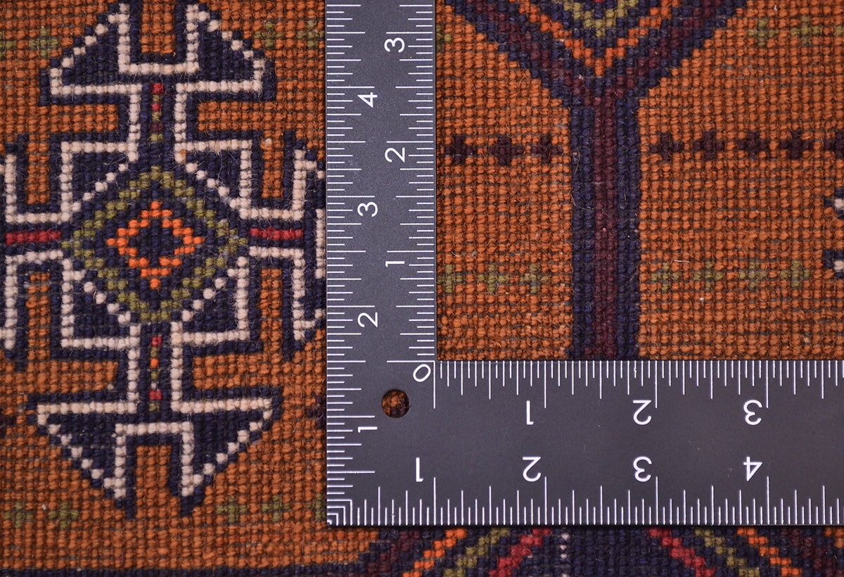 Close-up of the hand-knotted weave of a Old handmade Belouch mat/ small rug with a geometric pattern in fawn-brown, dark brown, cream, and small green accents, shown with a metal ruler to show scale of the knotting.