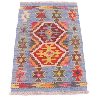 A Afghan Kilim mat handwoven with a geometric tribal pattern, featuring a dusty blue background with red, orange, yellow, and green motifs, finished with fringed edges.