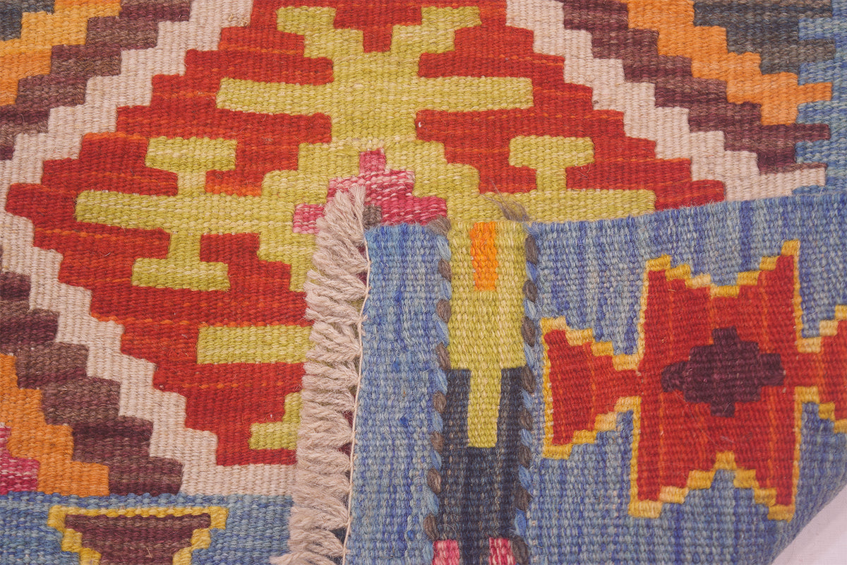 Close-up of a woven Afghan Kilim with a geometric pattern in red, yellow, blue, brown, and cream, showing a fringed edge.