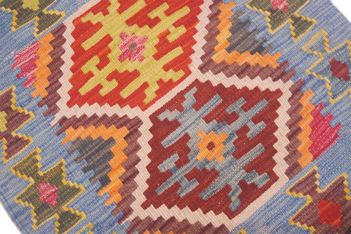 Close-up of a woven geometric Kilim rug with a multicolour pattern featuring red, yellow, blue, pink, and brown shapes on a light blue background.