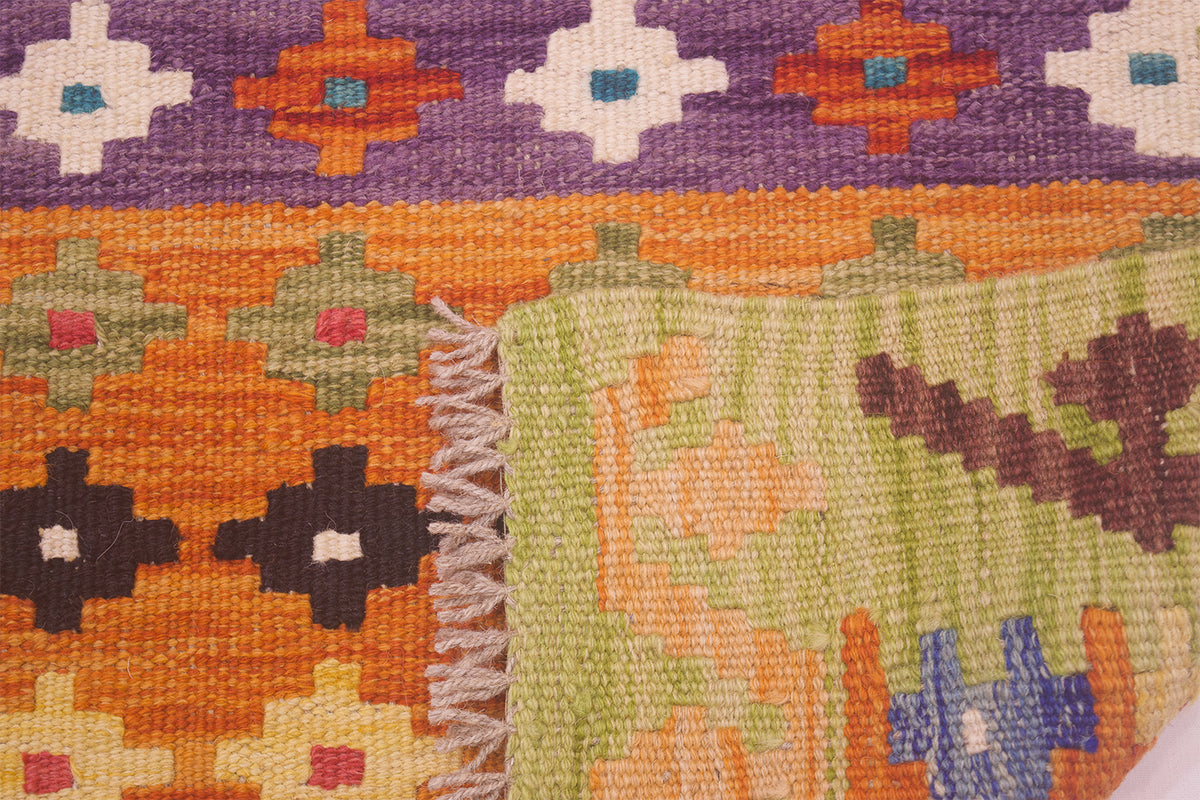 Close-up of a handwoven small Afghan Kilim with a multicolour geometric pattern, including purple, orange, green, brown, and blue sections with white fringe visible along one edge.