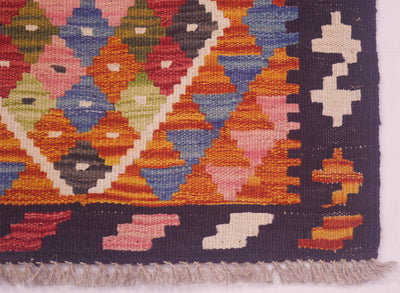 Close-up of a Small handwoven Afghan Kilim corner showing geometric diamond and triangle motifs in orange, blue, pink, green, red, and cream, with a dark navy border and light beige fringe.