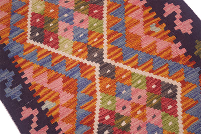 A close-up of a woven kilim rug featuring a vibrant geometric pattern with pink, orange, blue, green, and red shapes outlined in cream, bordered by a dark navy blue edge.