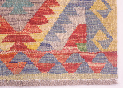 Close-up of a woven afghan kilim rug showing a geometric pattern with red, blue, yellow, beige, and grey shapes, finished with a fringed edge.