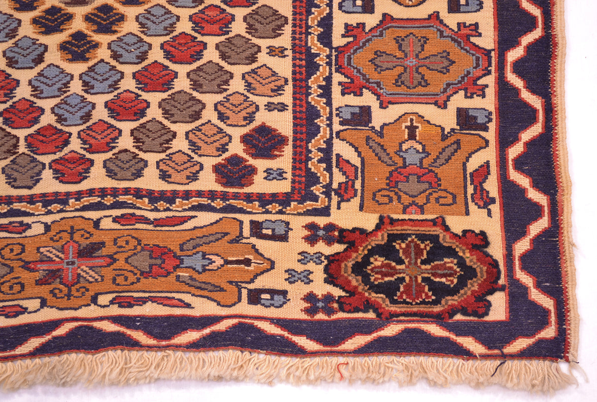 A close-up of a patterned Adraskan or Barjasta rug corner showing a beige base with navy, rust red, and brown geometric motifs and fringe along the bottom edge.