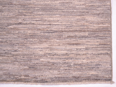A close-up of a corner of a textured handwoven plain rug with horizontal striations in taupe, beige, and grey tones, showing a frayed edge along the bottom.
