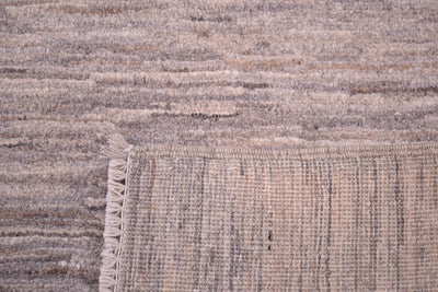 Close-up of a corner turned up of a handwoven plain Gabbeh rug corner showing a textured, fringed edge in neutral beige and taupe tones.