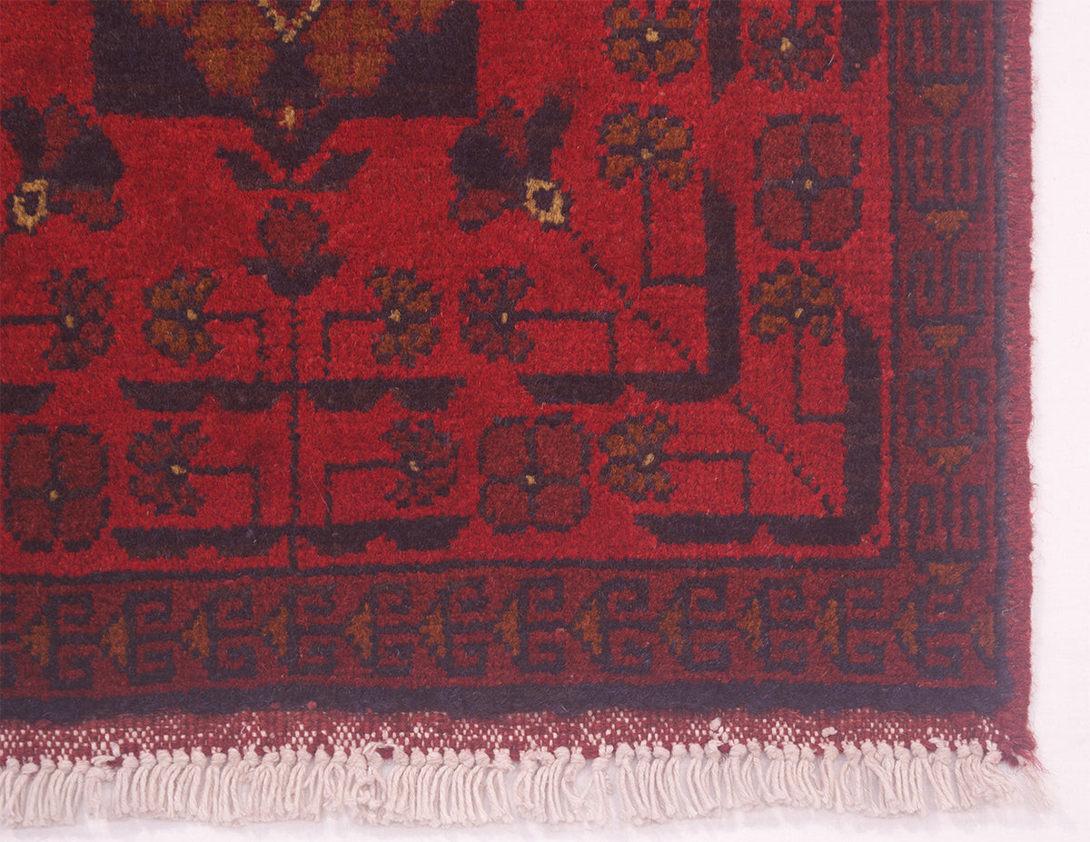A close-up of a red patterned Afghan runner rug corner showing floral motifs, a decorative border, and off-white fringe.