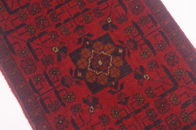 A close-up of a red runner rug with a central medallion and geometric floral motifs in dark brown, black, and muted gold.