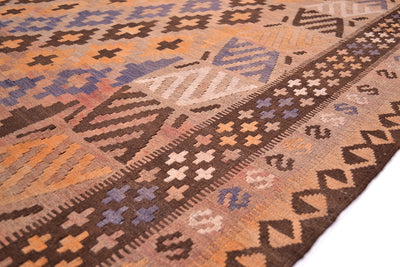 A close-up of a mature colour large old Kilim rug showing its detailed geometric patterns and border. The rug features a warm palette of brown, orange, beige, and muted blue, with a prominent dark brown patterned border on the right side.
