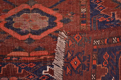 Close-up of the hand-knotted back corner turned up to show the weave of and Antique Ersari Chuval patterned rug showing a rust red field with dark blue geometric motifs and a fringed edge.