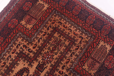 A close-up of a patterned antique Afghan Belouch prayer rug showing geometric and tribal-inspired motifs in deep red, terracotta, black, and beige.