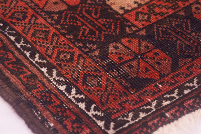 A close-up of the wear on a patterned Afghan Belouch antique rug with geometric tribal designs in red, black, and cream, showing the detailed woven texture and fringe on one side.
