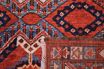 Close-up of a back corner turned up of an antique style Beshir patterned rug showing a red-orange field with navy and cream geometric motifs with white fringe visible at the lower left.