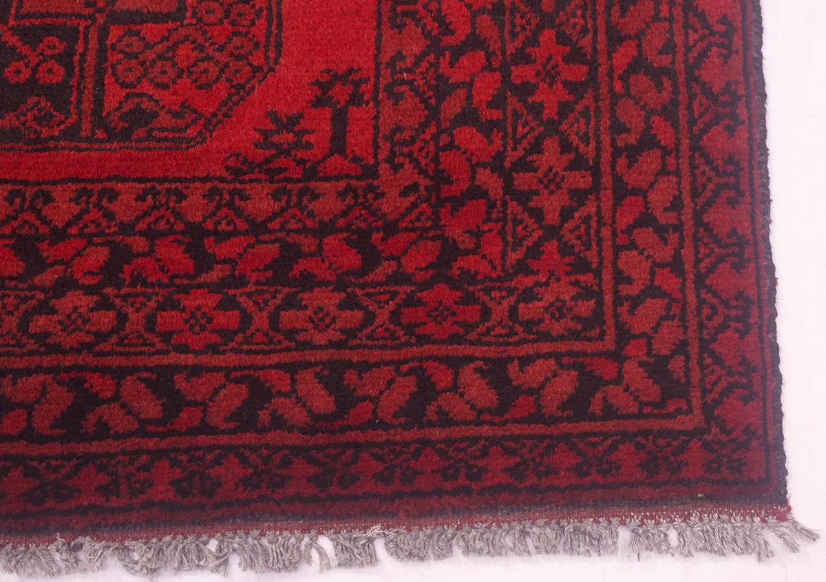 A close-up of a red and black Aqcha Afghan rug corner with grey fringe visible along the bottom edge, photographed against a white background.