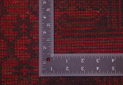 A close-up of the hand-knotted weave of a red and black Afghan Aqcha Fil Pai carpet with a dark L-shaped metal ruler placed on top to show the material texture and scale of the knot density. 