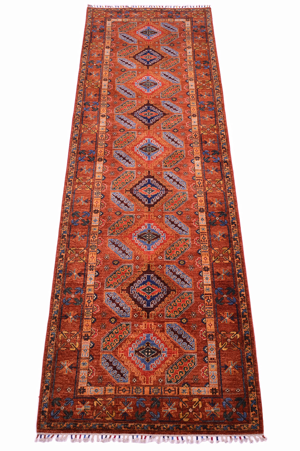 A long rectangular area runner with a traditional geometric pattern in rust red, orange, navy blue, beige, and cream, featuring a central field, decorative borders, and fringed ends.