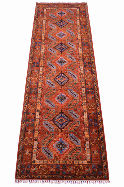 A long rectangular area runner with a traditional geometric pattern in rust red, orange, navy blue, beige, and cream, featuring a central field, decorative borders, and fringed ends.
