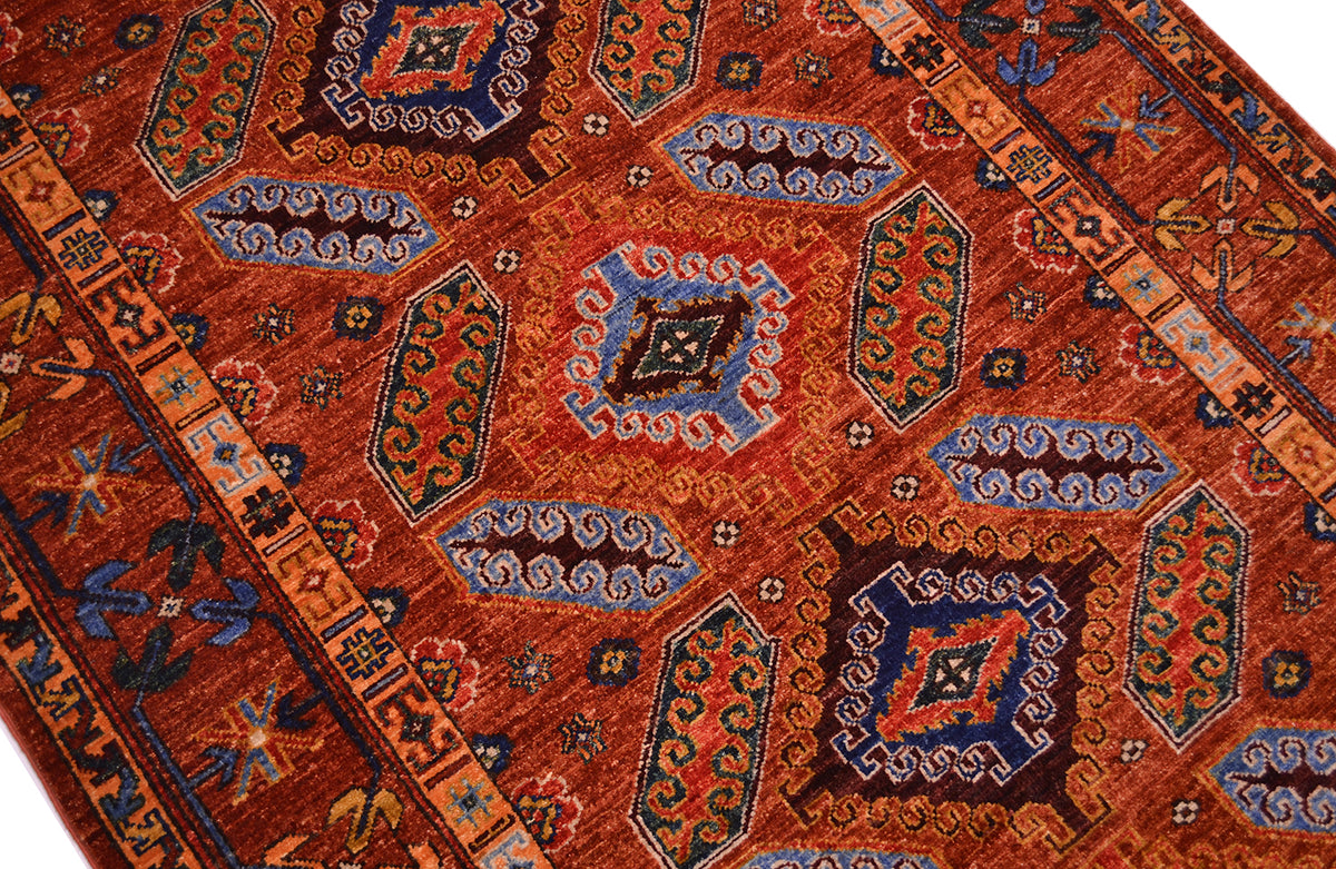 A close-up of a traditional patterned rug with a warm cinnamon field, featuring geometric medallions and borders in blue, navy, beige, and brown.