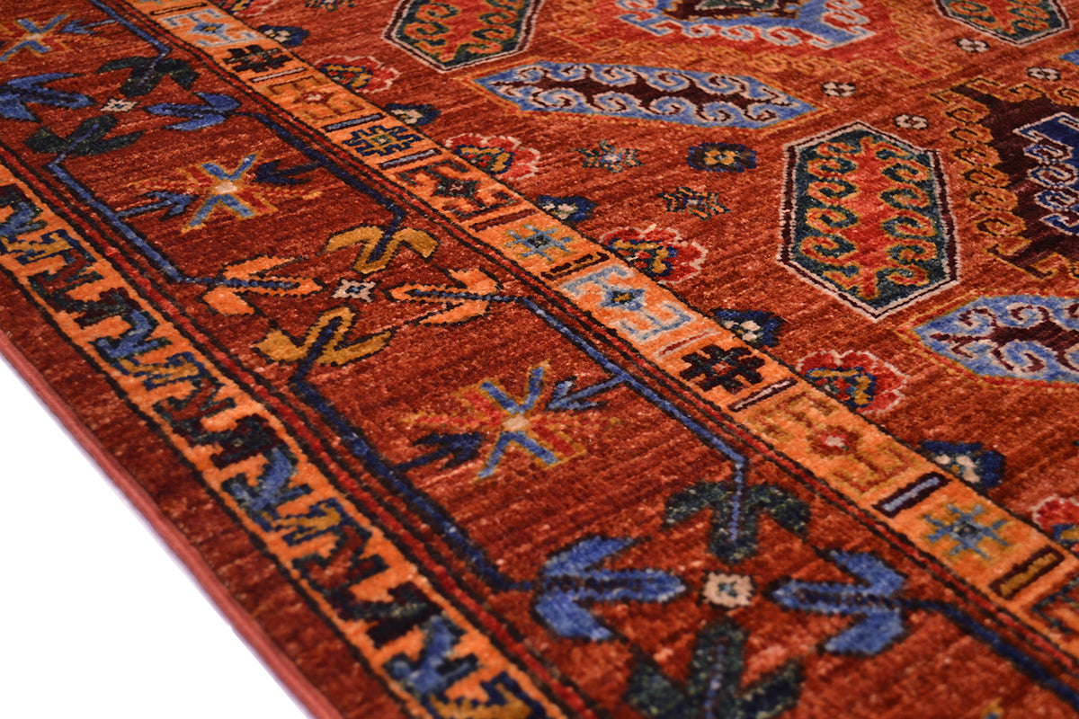 A close-up of a traditional patterned rug with a warm cinnamon red field, blue and orange geometric borders, and floral motifs.