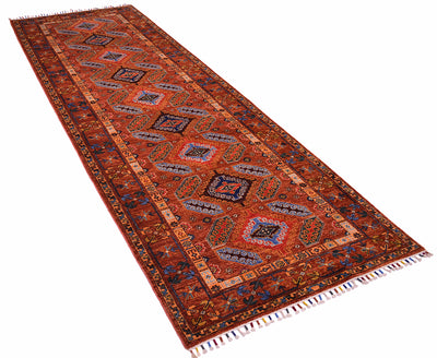 A long rectangular area rug with a traditional geometric pattern in cinnamon red, orange, navy, beige, and blue, featuring fringed ends, shown on a plain white background.