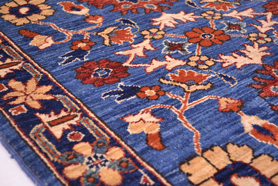 A close-up of a traditional patterned Afghan Aryana rug with a medium blue field, featuring floral motifs in rust red, beige, and orange, and a matching blue border.