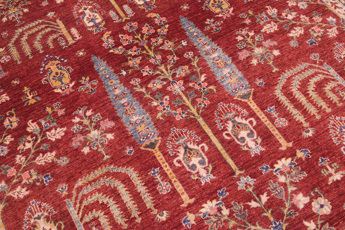 A close-up of a traditional patterned pictorial Afghan Aryana handmade carpet with a deep red background, featuring ornate floral motifs and stylised tree-like designs in beige, blue, and gold.