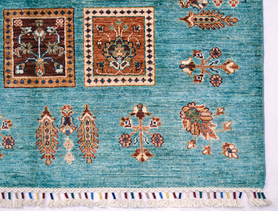 A close-up of a corner of a handmade Afghan Bakhtiari design turquoise carpet with traditional geometric and floral motifs in brown, beige, and cream, finished with a fringed edge.