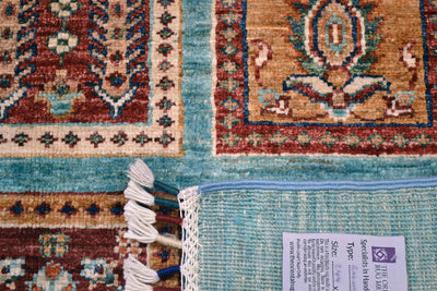 Close-up of a corner turned up showing the hand-knotted weave of an Afghan hand woven Bakhtiari design turquoise carpet showing a turquoise blue field with cream fringe, a red and beige patterned section, and a grey product label attached near the edge.