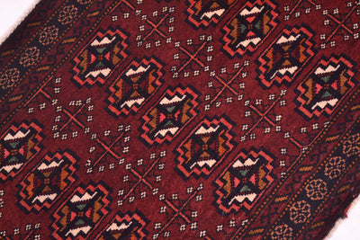 A close-up of a patterned Afghan Belouch small rug with a deep maroon field, featuring repeating geometric motifs in black, beige, orange, and green. 